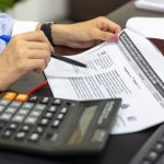 Pessoa analisa documentos financeiros com calculadora e caneta sobre uma mesa.