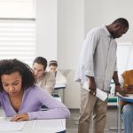 Estudantes de diferentes perfis realizam uma prova em uma sala de aula moderna, sob supervisão de uma professora.