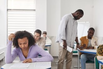 Estudantes de diferentes perfis realizam uma prova em uma sala de aula moderna, sob supervisão de uma professora.