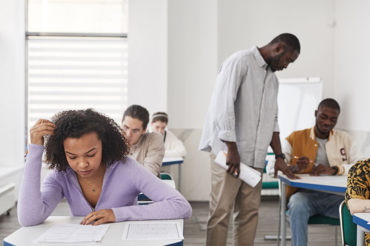 Estudantes de diferentes perfis realizam uma prova em uma sala de aula moderna, sob supervisão de uma professora.