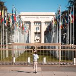 Pessoa fotografa o escritório da ONU em Genebra, diante de bandeiras internacionais.